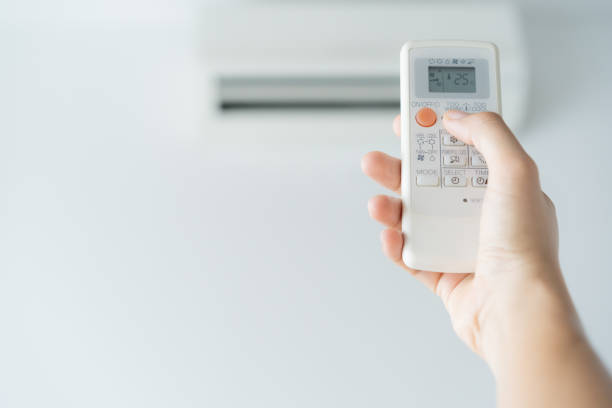 Hand holding a remote control pointed toward a wall-mounted split air conditioner, adjusting the temperature settings in a modern indoor environment.